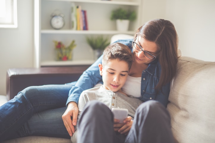 Photography son lying on the sofa and holding his hand smart-phone, the mother sits and looks at his smart phone