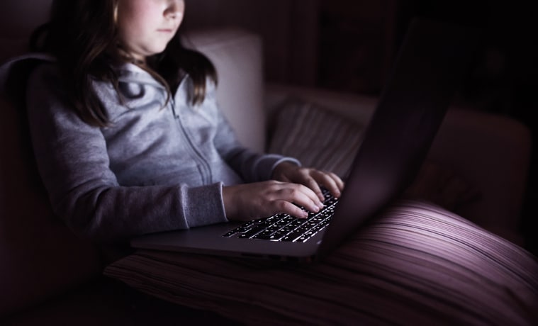 Unrecognizable girl, sitting in a dark, playing with laptop. Child at home, sitting on sofa.